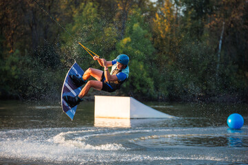 An athlete does a trick from a springboard. A rider jumps on a wakeboard against a background of a green forest. Sunset on the lake