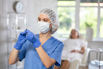 Female medical person wearing blue uniform, facial mask and bouffant cap standing at examination room with syringe in her hands