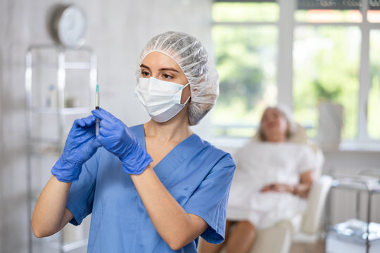 Female medical person wearing blue uniform, facial mask and bouffant cap standing at examination room with syringe in her hands