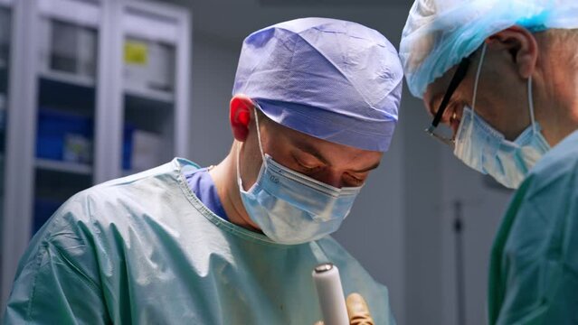 Younger male surgeon holding a chisel at operation. Adult doctor uses a mallet and hits by the chisel.