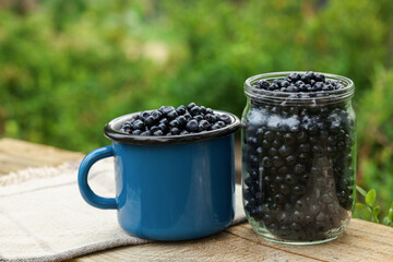 Jar and cup of delicious bilberries on wooden table outdoors
