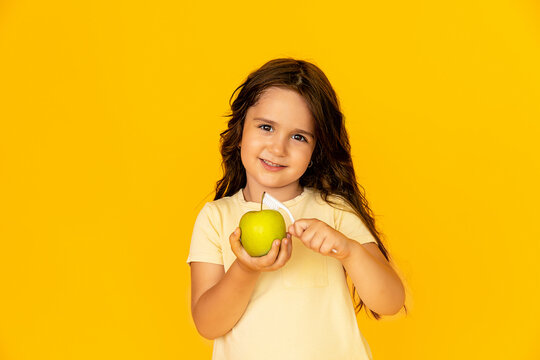 Little girl in yellow t-shirt smiling and peeling a green apple with a toothbrush on yellow studio background. Dental care and health. Pediatric dentistry concept. Mock up. Space for text.