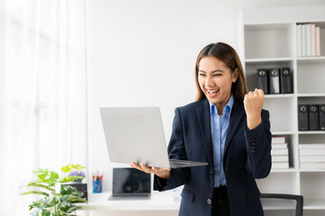 Young Asian business woman wearing suit and using laptop on gray baclground Cheerful young woman celebrating her achievement while reading good news on laptop from home office