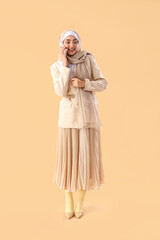Young Muslim woman in stylish suit talking by mobile phone on beige background