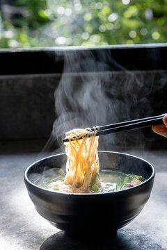 Hand uses chopsticks to pickup tasty noodles with steam and smoke in bowl on wooden background, selective focus. Asian meal on a table, junk food concept