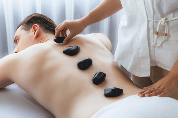 Hot stone massage at spa salon in luxury resort with day light serenity ambient, blissful man customer enjoying spa basalt stone massage glide over body with soothing warmth. Quiescent