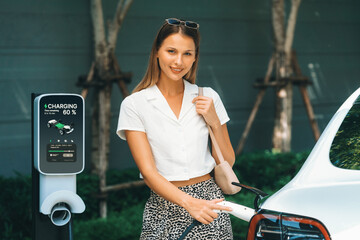 Young woman travel with EV electric car charging in green sustainable city outdoor garden in...