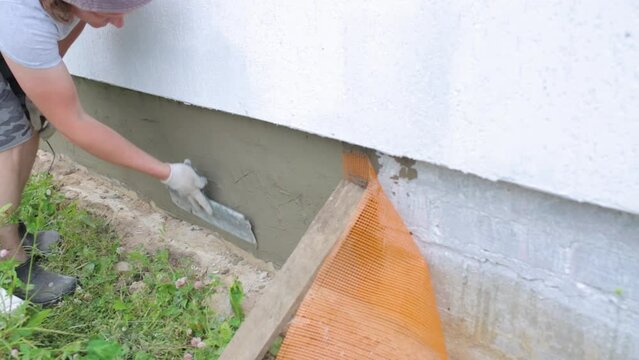 A worker uses a large trowel to putty the foundation of a house. Covering the foundation with mesh and putty. A man plasters a house and insulates it