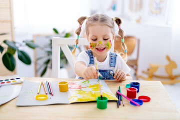 a little child girl paints a drawing in an album and smiles with a dirty face and hands, a happy child draws a picture at home at the table