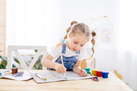 a little child girl paints a drawing with a brush in an album and smiles, a happy child draws a gouache picture at home at the table
