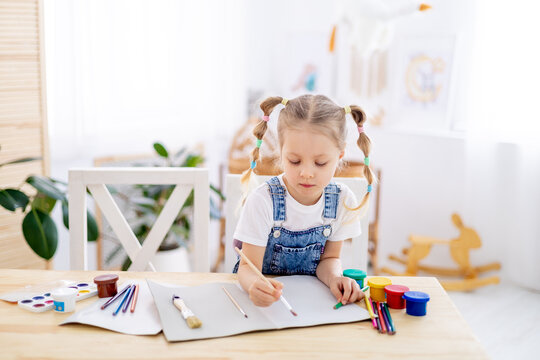 a little child girl paints a drawing with a brush in an album and smiles, a happy child draws a gouache picture at home at the table