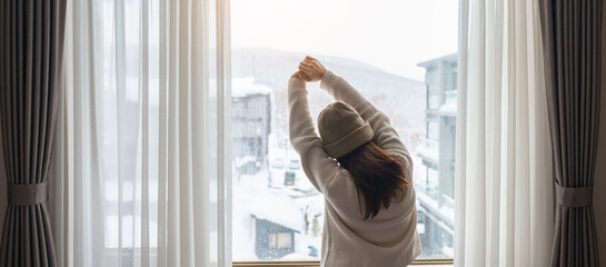 young woman in sweater looking through the window in winter season, happy female rising arms and...