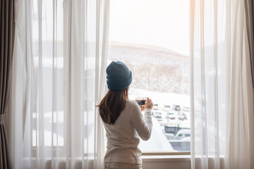 young woman in sweater with cup of coffee looking through the window in winter season, happy female enjoying snowfall outdoor view at apartment or home in the morning. Waking and Relaxing concept