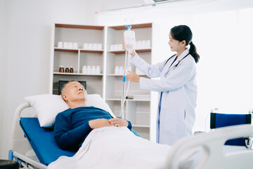 Asian doctor in white suit take notes while discussing and Asian elderly, man patient who lying on bed with receiving saline solution in hospital .