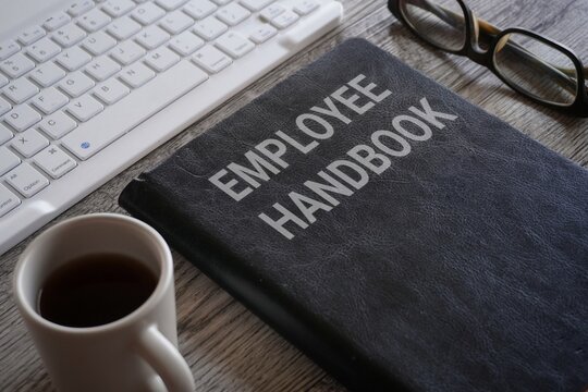Closeup image of book with text EMPLOYEE HANDBOOK surrounded by cup of coffee and glasses on office desk