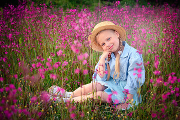 Portrait of a blonde little girl in a straw hat and blue clothes in pink meadow flowers