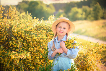 Portrait of a blonde little girl in a straw hat and blue clothes against a yellow bush in spring