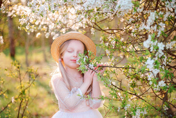 Portrait of a blonde little girl in a straw hat and pink dress against a background of flowering trees in spring