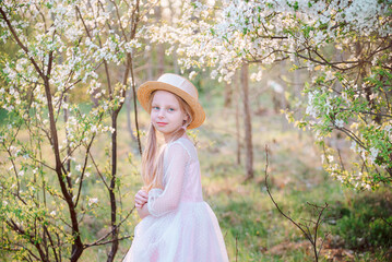 Portrait of a blonde little girl in a straw hat and pink dress against a background of flowering trees in spring