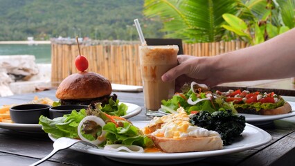 Man drink iced cocktail, enjoying vegan food by ocean. Close-up menu of vegan food in cafe poached eggs, salad, healthy burger, avocado toast, cocktail. Concept visit cafe to enjoy healthy vegan food.