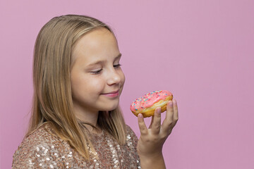 Cheerful kid girl biting donut and having fun against pink color studio wall background. Colorful holiday birthday party concept. Positive emotion.