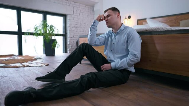 Wide shot stressed overburdened young businessman sitting on floor leaning on bed thinking. Side angle view portrait of tired worried Caucasian man looking away planning business startup