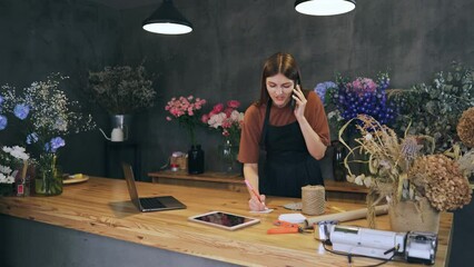 Young beautiful woman florist in an apron in a flower shop receives an order by phone to make a bouquet