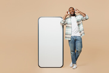 Full body young man he wear light shirt casual clothes big huge blank screen mobile cell phone with workspace copy space mockup area listen to music in headphones isolated on plain beige background.