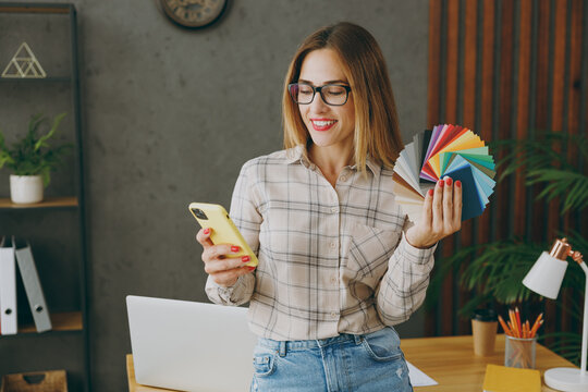Young happy designer employee business woman wear shirt casual clothes glasses sit work at office desk with pc laptop hold color palette use mobile cell phone internet. Achievement career job concept.