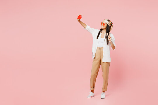 Full body young latin woman wear white shirt casual clothes sunglasses doing selfie shot pov on mobile cell phone show v-sign isolated on plain pastel light pink background studio. Lifestyle concept.