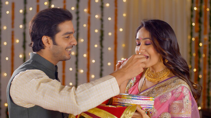Young attractive couple preforming Karwa Chauth traditions - Indian festival. Handsome husband offering sweets to her pretty wife to complete her fast successfully on the occasion of Karwa Chauth