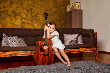 Cute little child with guitar in hands at living room, pensive looking. Elegant adorable kid girl in white dress with guitar sitting on couch at home. Leisure music hobby concept. Copy ad text space