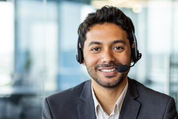 Close-up portrait of young successful businessman with headset phone, customer support worker in business suit smiling and looking at camera, workplace inside office. - obrazy, fototapety, plakaty