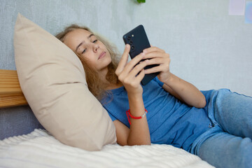 A cute teenage girl watches videos or writes messages on her smartphone while lying on the bed in the bedroom. Communication of modern children