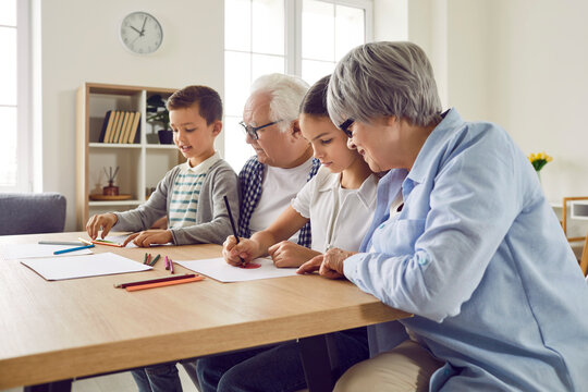Senior couple grandparents spending time with their grandchildren boy and girl at home on weekend drawing with colored pencils together sitting at the table. Family leisure concept.