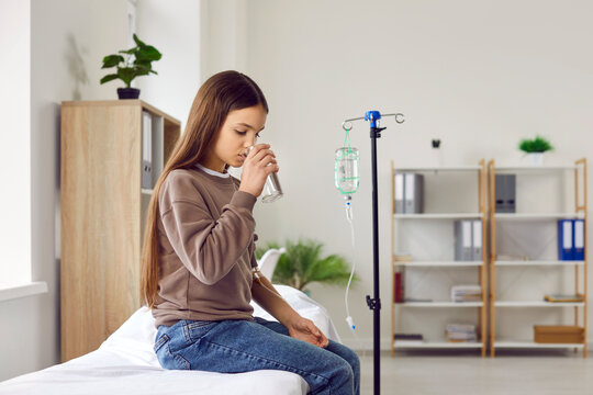 Child patient getting intravenous therapy at clinic or hospital. Sick girl with IV line venous catheter in arm sitting on medical bed, taking medicine and drinking water. Health care treatment concept