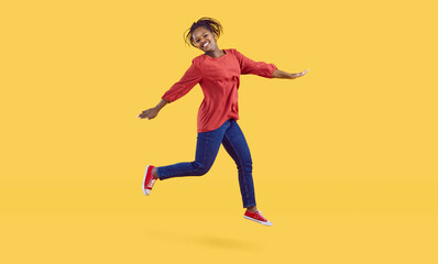 Fototapeta na wymiar Happy young black woman having lots of fun in the studio. Cheerful carefree African American girl wearing comfortable blue jeans jumping high in the air isolated on a vibrant yellow colour background