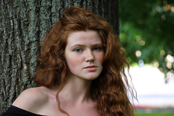 Portrait of ginger girl with long red hair and freckles standing near the tree in park. Young sensual model, natural female beauty	