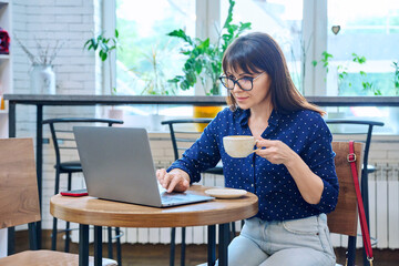 Middle-aged woman drinking coffee looking at laptop screen, sitting in coffee shop