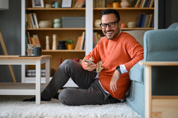 Happy handsome freelancer texting over smart phone while sitting by sofa on carpet in home office