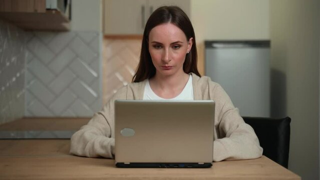 Portrait of a cute positive girl, sitting at home in the kitchen with a laptop, working or studying remotely, looking at the camera, smiling amiably.