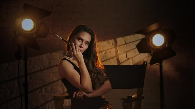Woman in black dress sitting on a director's chair, holding cigarette holder and smoking, looking at the camera. Slow motion.