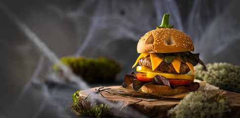 Halloween Burger banner. Halloween party creative burger concept with big beef patties with pumpkin head rolls, tomato and pepper on wooden board on dark background with moss. Copy space