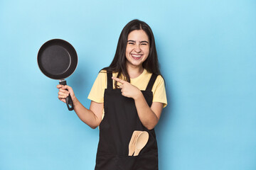 Obraz premium Young woman with pan on blue studio smiling and pointing aside, showing something at blank space.