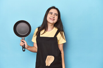 Young woman with pan on blue studio dreaming of achieving goals and purposes