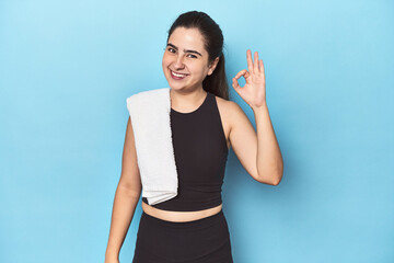 Sporty woman with towel on blue studio cheerful and confident showing ok gesture.