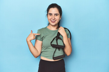 Young woman with resistance bands on blue person pointing by hand to a shirt copy space, proud and confident