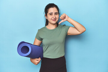 Sporty woman with yoga mat on blue showing a dislike gesture, thumbs down. Disagreement concept.