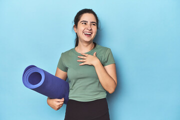 Sporty woman with yoga mat on blue laughs out loudly keeping hand on chest.