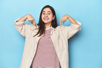 Young Caucasian woman on blue backdrop points down with fingers, positive feeling.